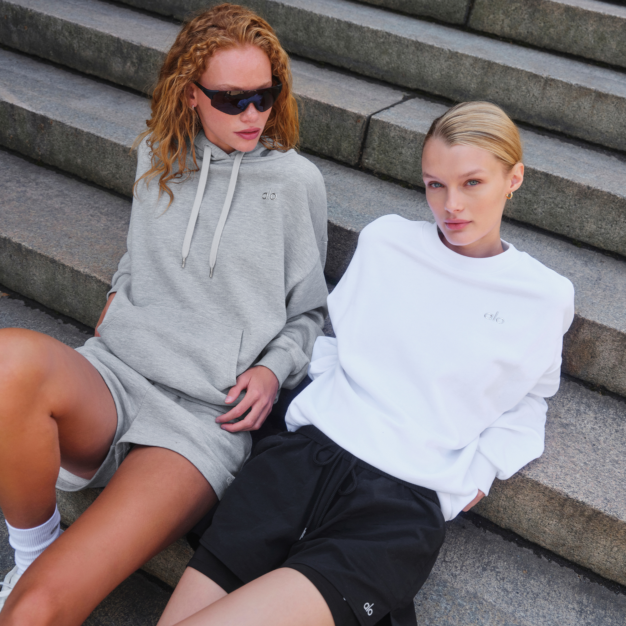 Two women in casual hoodies and shorts sitting on stone steps