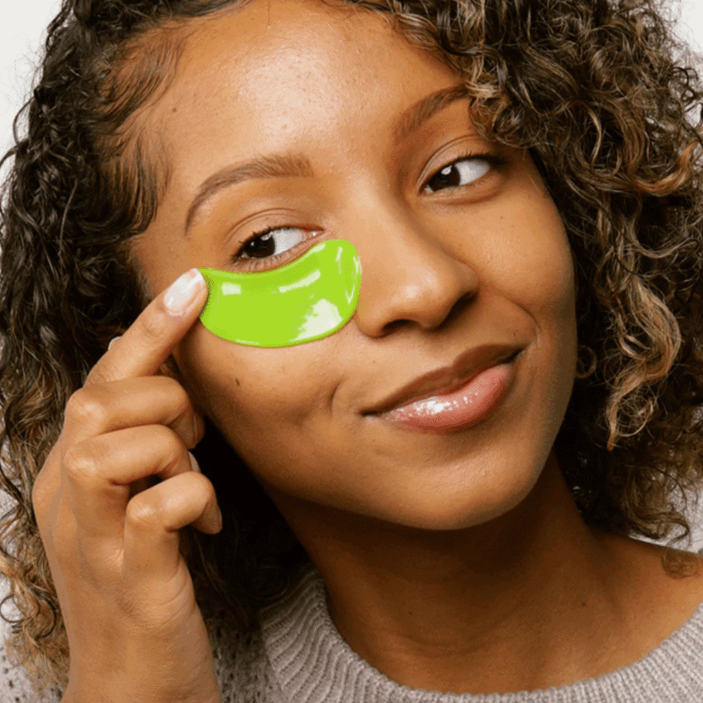 Close-up of a woman with curly hair applying a bright green under-eye gel patch while smiling gently and looking sideways, wearing a knitted top.