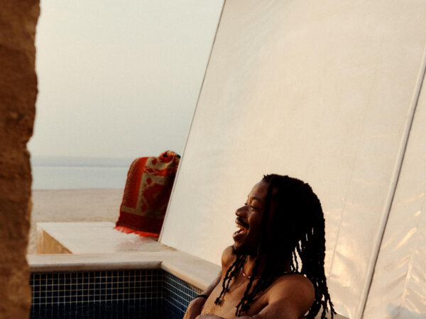 Man with long dreadlocks relaxing and laughing while partially submerged in a tiled outdoor pool near a beach, with a white tent-like structure and a red blanket in the background.