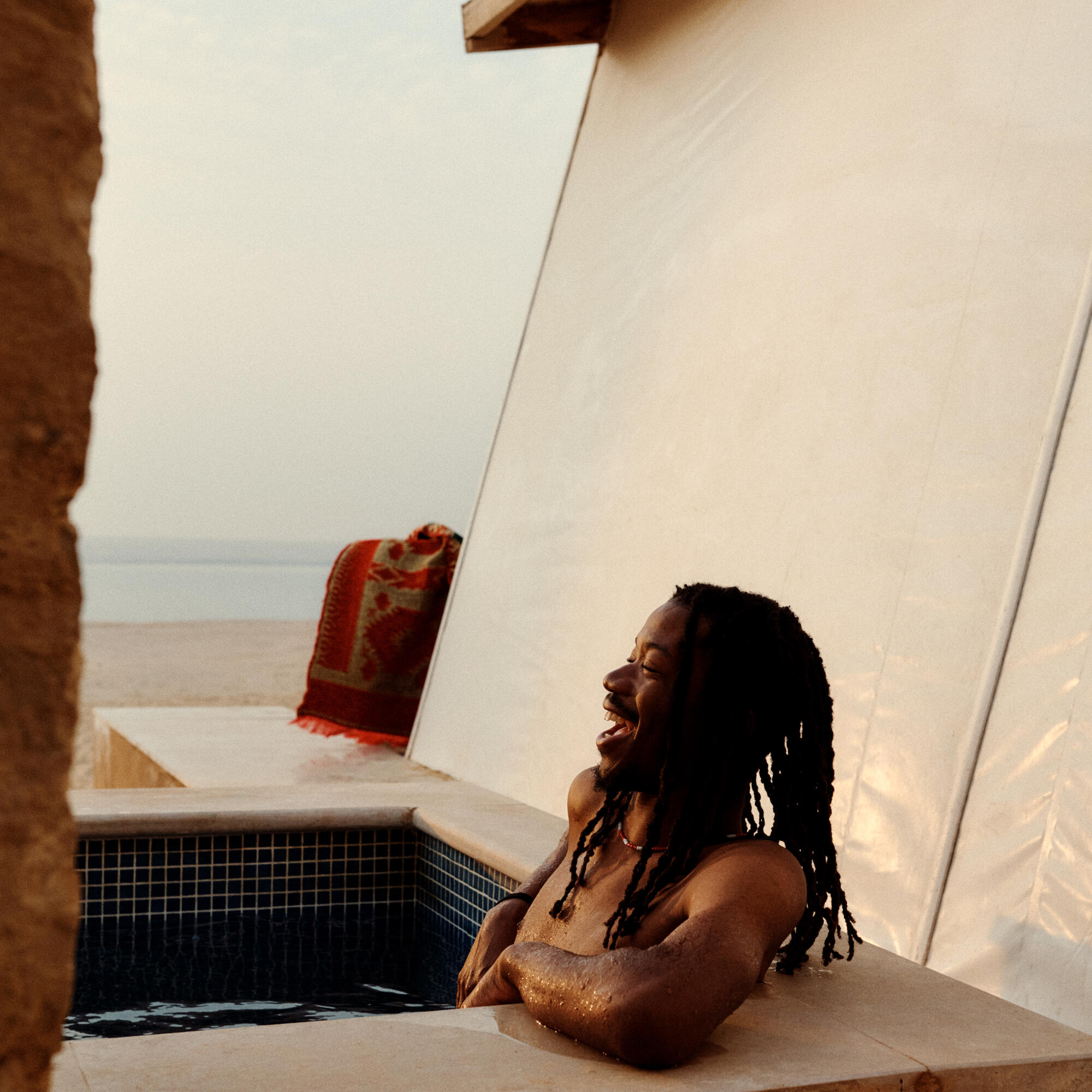 Man with long dreadlocks relaxing and laughing while partially submerged in a tiled outdoor pool near a beach, with a white tent-like structure and a red blanket in the background.