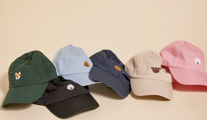 Six baseball caps in different colors arranged in a row on a beige background, each featuring a small embroidered dog face on the front, including green, black, light blue, navy, beige, and pink caps.