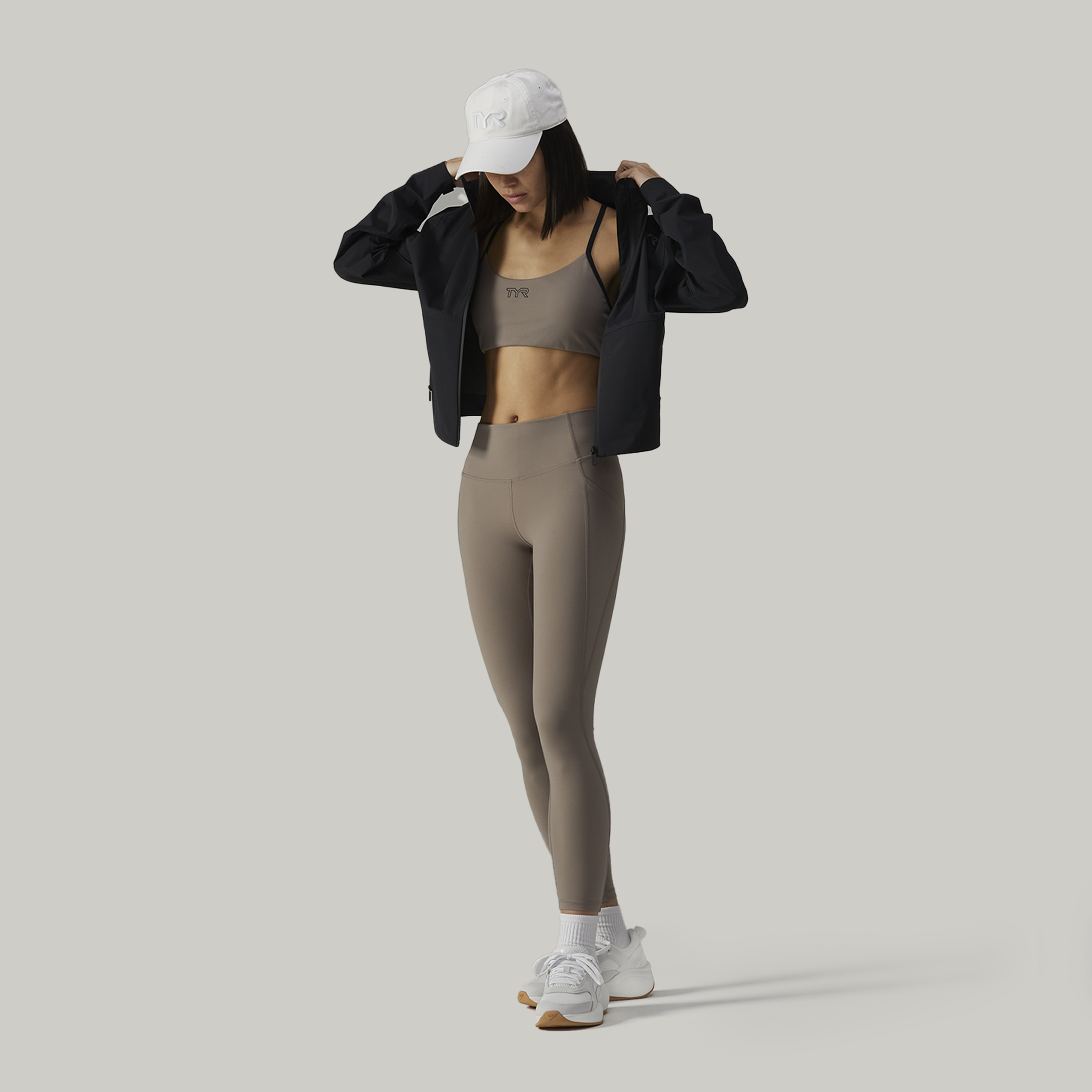 Young woman wearing a white cap, black jacket, taupe sports bra, and matching high-waisted leggings, posing against a plain light grey background.