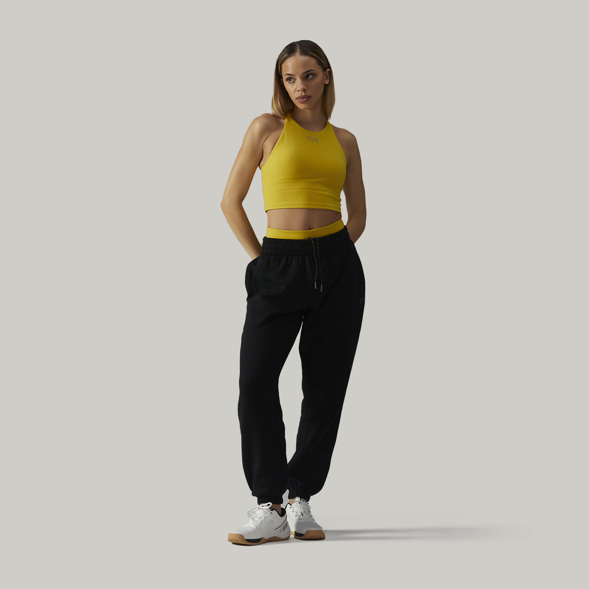 Young woman standing with hands in pockets wearing a yellow sleeveless crop top, black sweatpants, and white sneakers against a plain light grey background