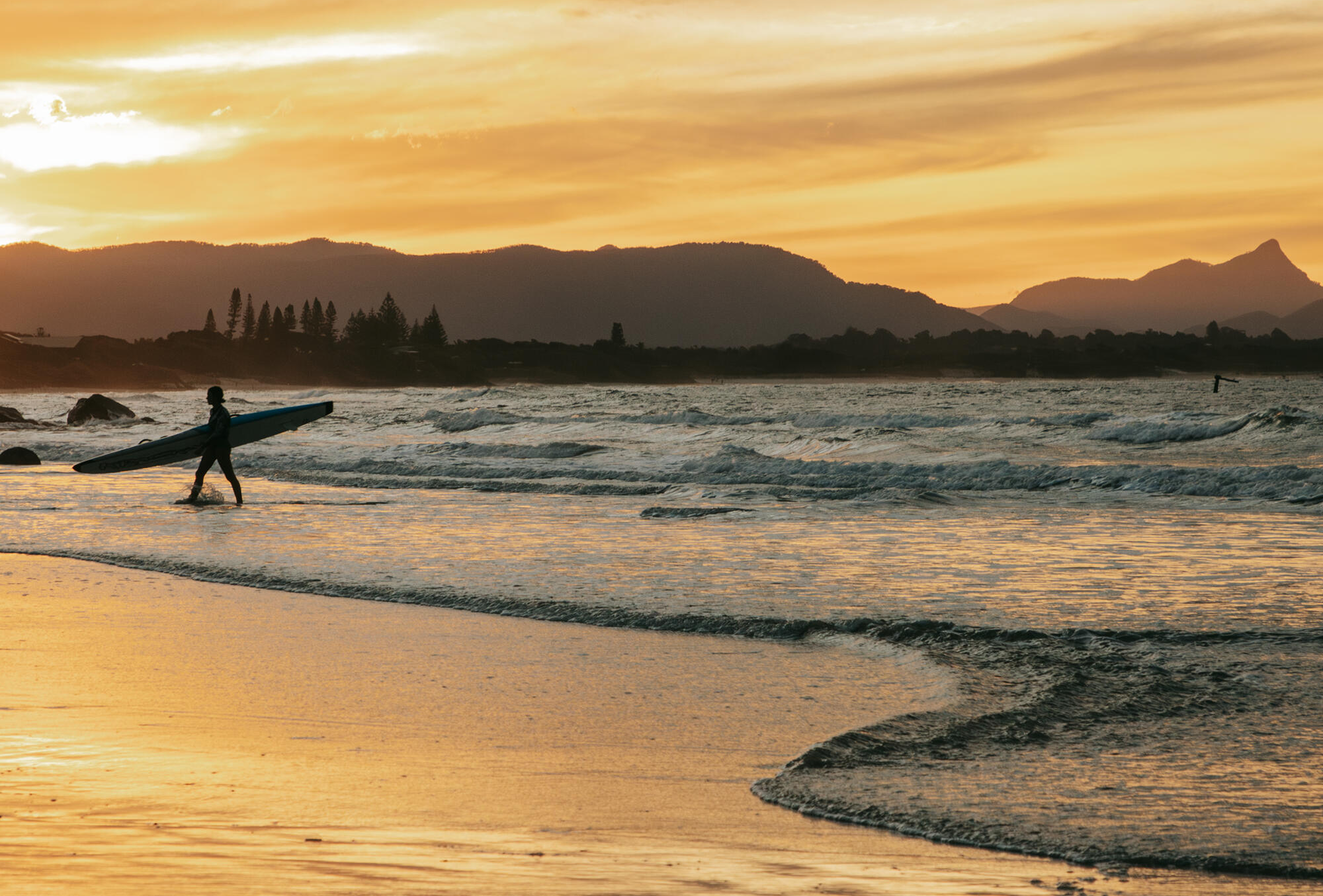 Silhouette of a person carrying a surfboard walking along a beach at sunset with waves rolling in and mountains in the background.