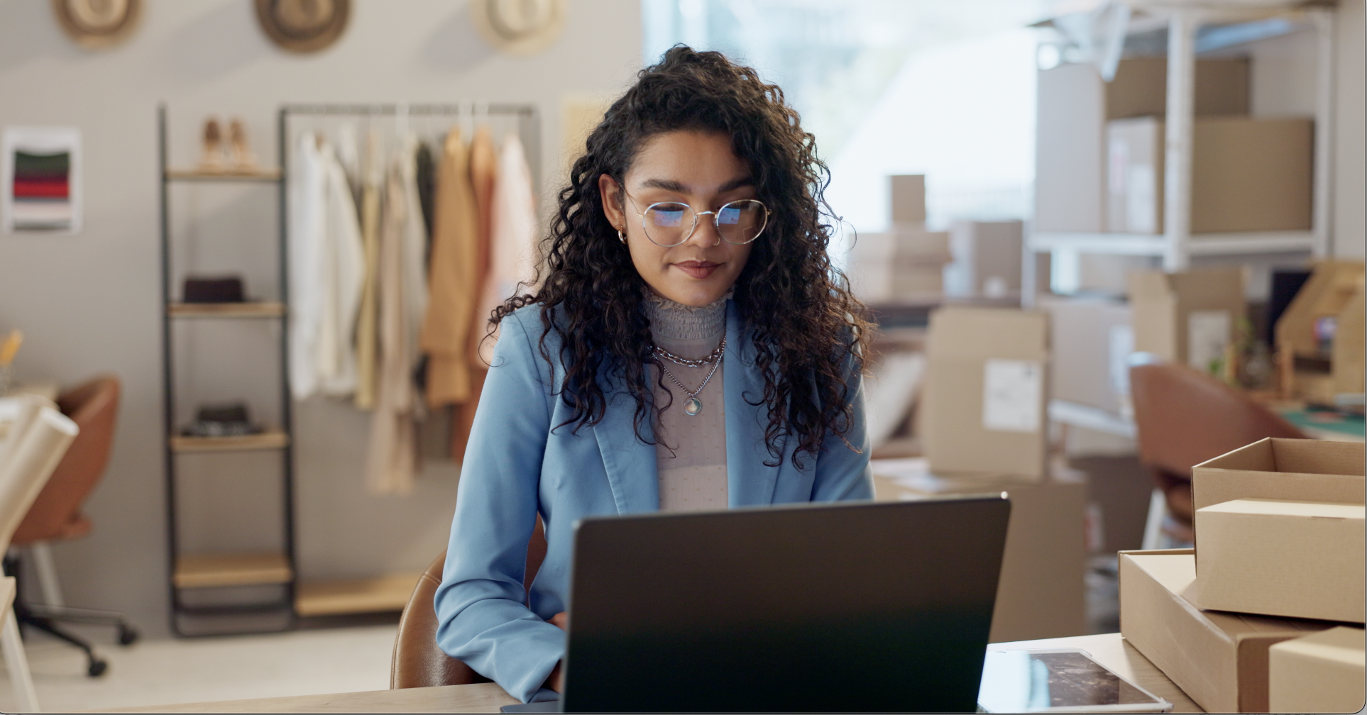 A woman with curly hair and glasses wearing a blue blazer is working on a laptop in an office filled with cardboard boxes and clothing racks in the background.