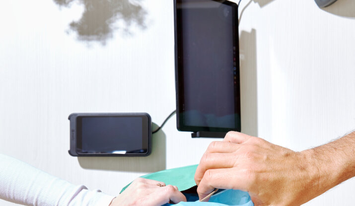 Two hands exchanging a small gift wrapped in blue tissue paper over a white table with electronic devices and a vase of flowers casting a shadow.