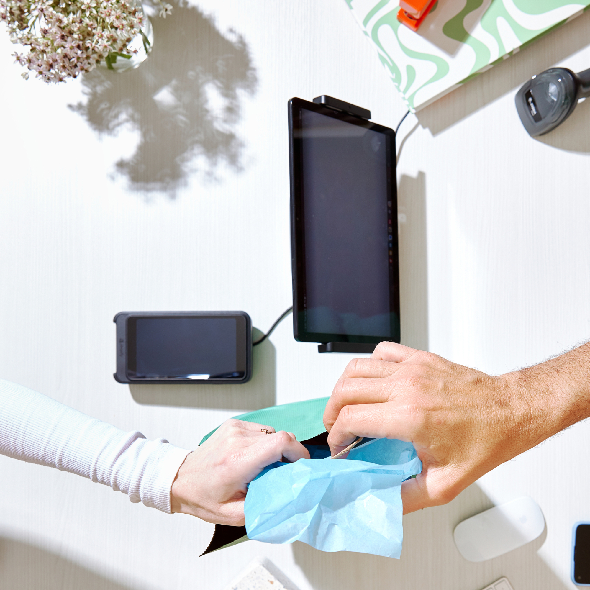 Two hands exchanging a small gift wrapped in blue tissue paper over a white table with electronic devices and a vase of flowers casting a shadow.
