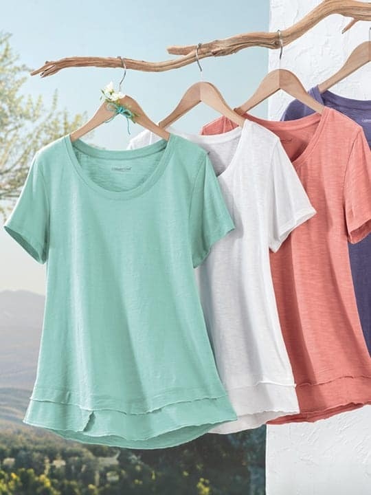 Four short-sleeve t-shirts in mint green, white, coral, and purple hanging on wooden hangers from a natural wooden branch with a small white flower decoration, outdoors against a clear sky and landscape background.