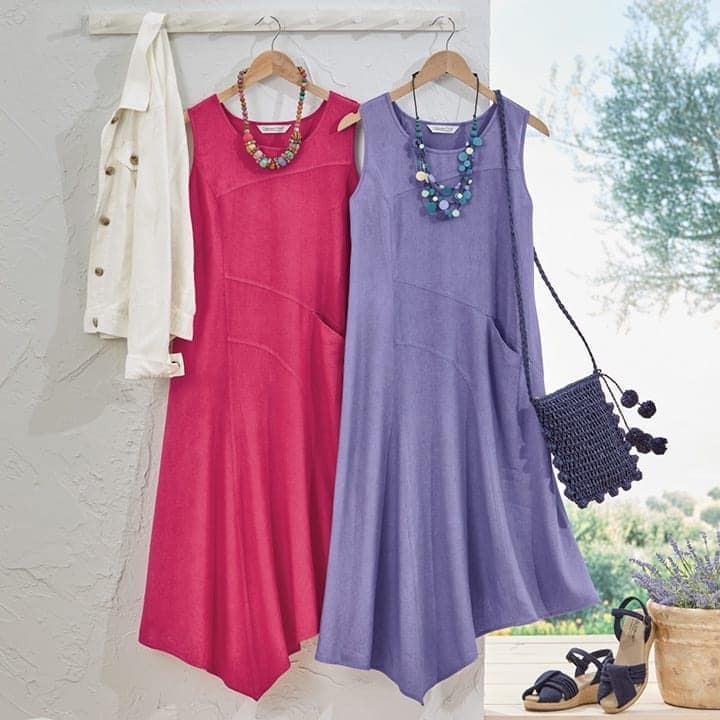 Two sleeveless dresses hanging on wooden hangers against a light textured wall, one in bright pink paired with a colorful beaded necklace and a cream jacket, the other in lavender with a blue beaded necklace and a navy woven shoulder bag, next to a pair of navy sandals and a potted plant on the floor