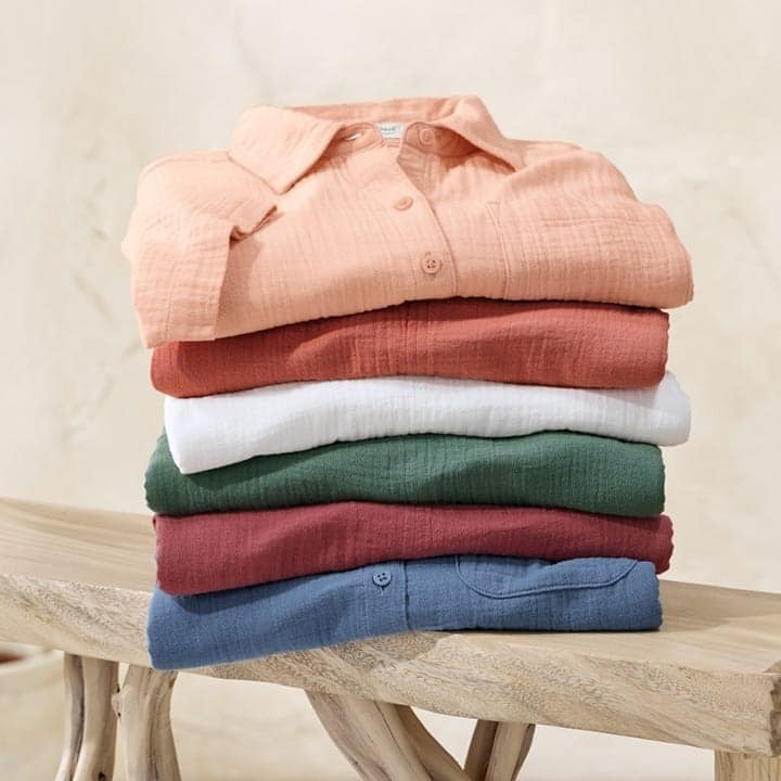 Stack of six neatly folded button-up shirts in various colors including peach, rust, white, green, burgundy, and blue on a wooden bench against a neutral background.