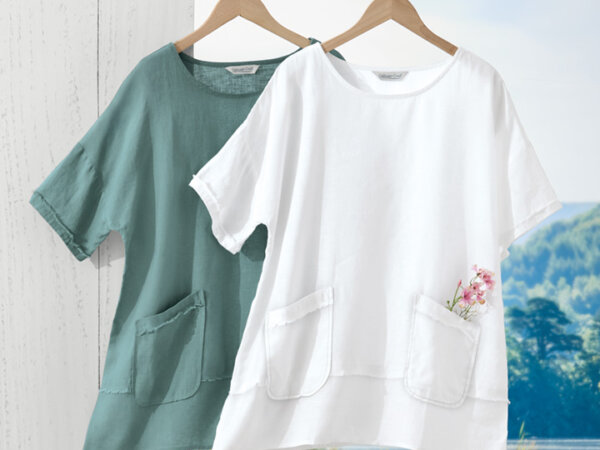 Two linen short-sleeve tops hanging on wooden hangers, one in white with a small pink flower in the pocket and the other in muted green, against a white wooden background and a scenic outdoor view.