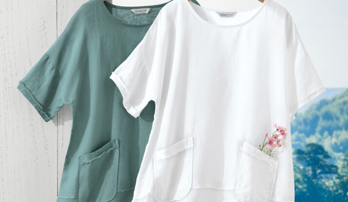 Two linen short-sleeve tops hanging on wooden hangers, one in white with a small pink flower in the pocket and the other in muted green, against a white wooden background and a scenic outdoor view.