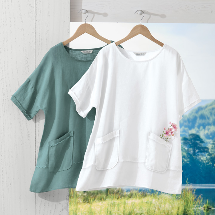Two linen short-sleeve tops hanging on wooden hangers, one in white with a small pink flower in the pocket and the other in muted green, against a white wooden background and a scenic outdoor view.