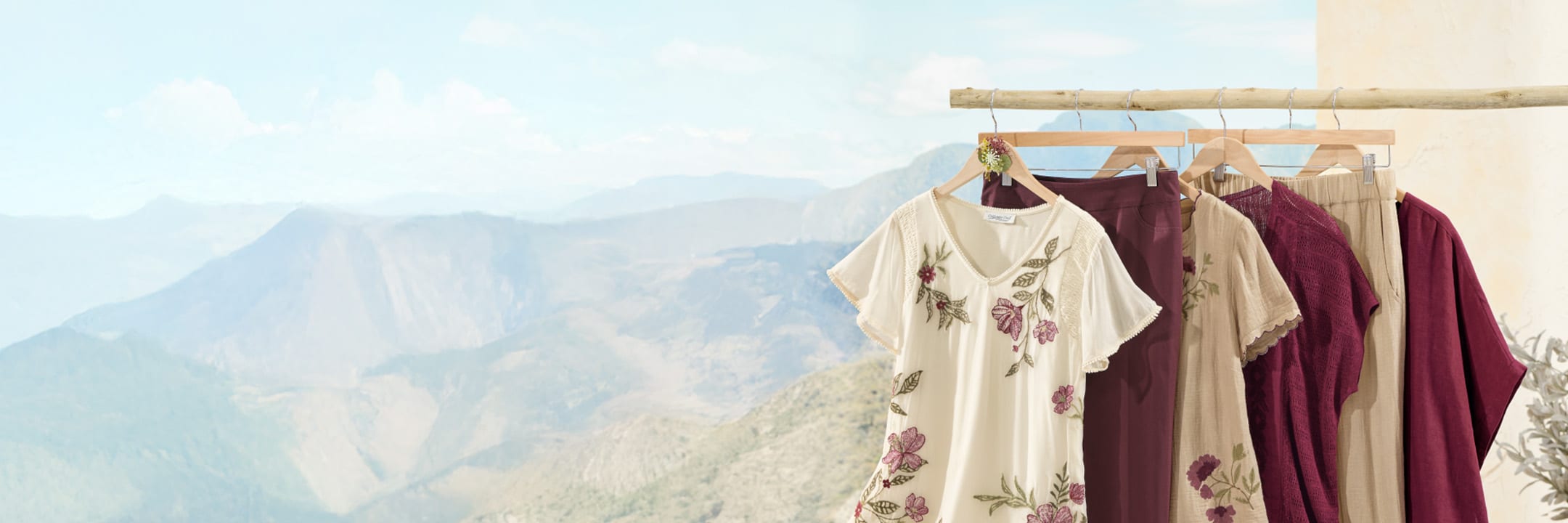 A selection of women's clothing hanging on wooden hangers from a rustic wooden rod against a mountainous outdoor background, featuring a cream embroidered blouse and various tops and pants in shades of burgundy and beige.