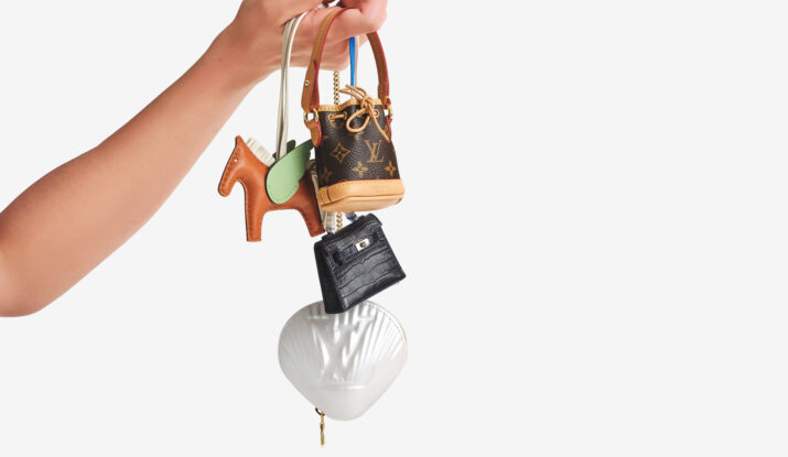 Hand with dark nail polish holding four small designer bag charms including a brown horse with green wings, a Louis Vuitton bucket bag, a black crocodile-patterned bag, and a white shell-shaped bag with embossed logo on a white background.
