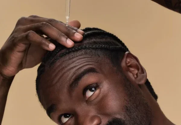 Close-up of a man with braided hair applying oil or serum to his scalp using a dropper against a plain beige background