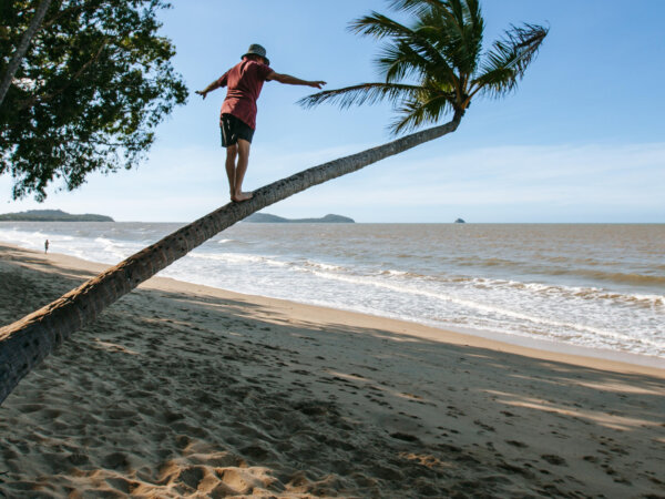 Person balancing and walking barefoot on a leaning palm tree trunk at a sandy beach with ocean waves and distant islands under a clear blue sky