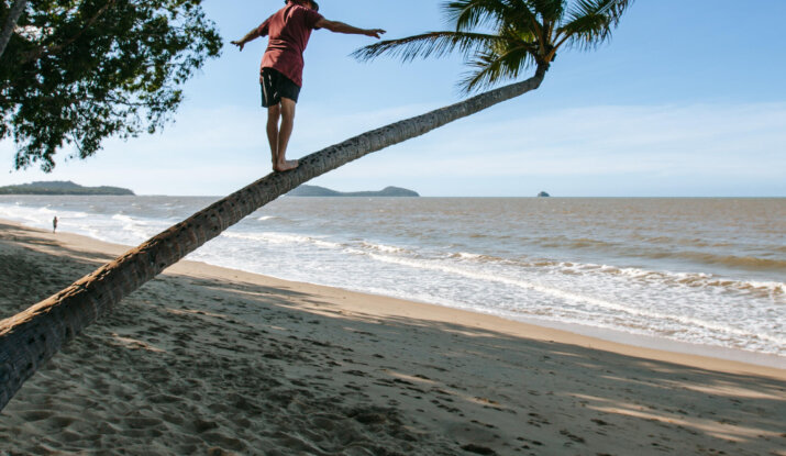 Person balancing and walking barefoot on a leaning palm tree trunk at a sandy beach with ocean waves and distant islands under a clear blue sky