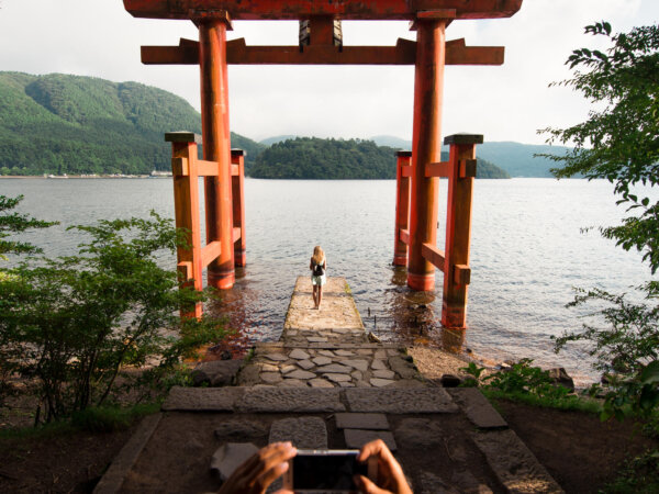 A person taking a photo with a smartphone of another person standing at the base of a large red torii gate by a lake with forested hills in the background under a cloudy sky.