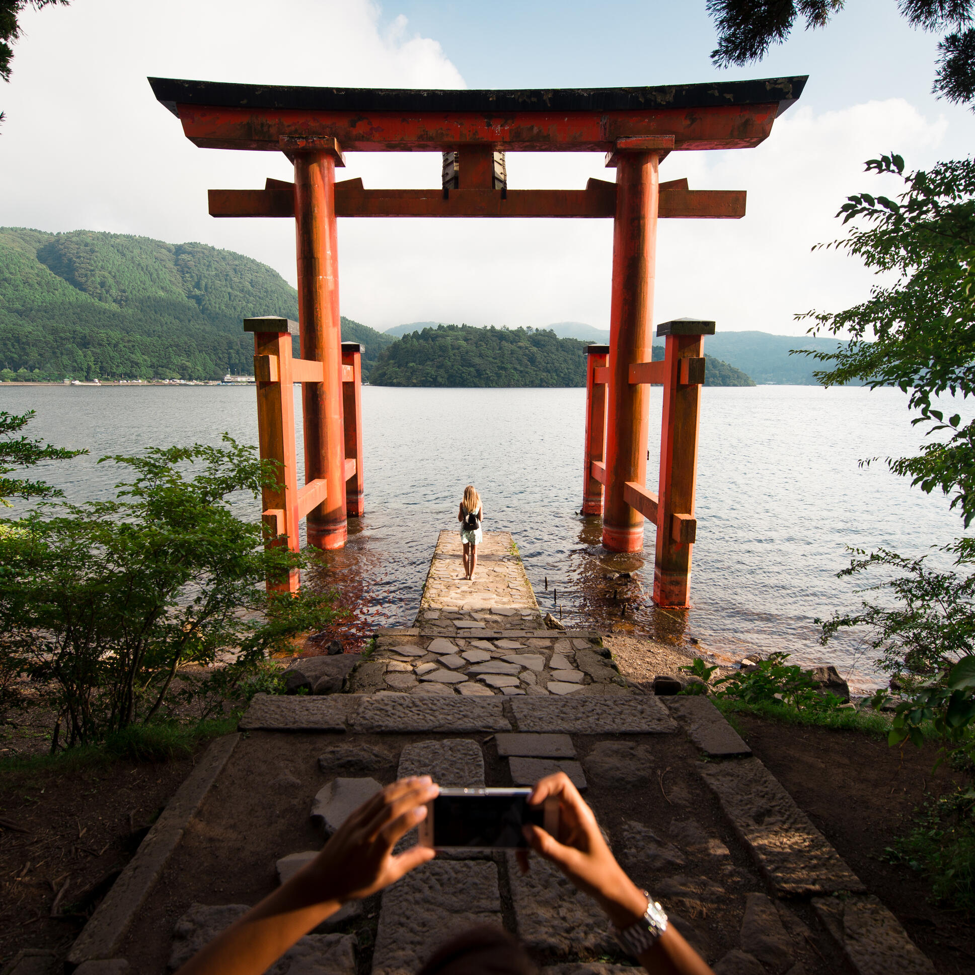 A person taking a photo with a smartphone of another person standing at the base of a large red torii gate by a lake with forested hills in the background under a cloudy sky.