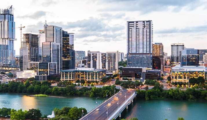 Aerial view of a city skyline with modern high-rise buildings, a bridge crossing a river, and green park areas under a cloudy sky at dusk.