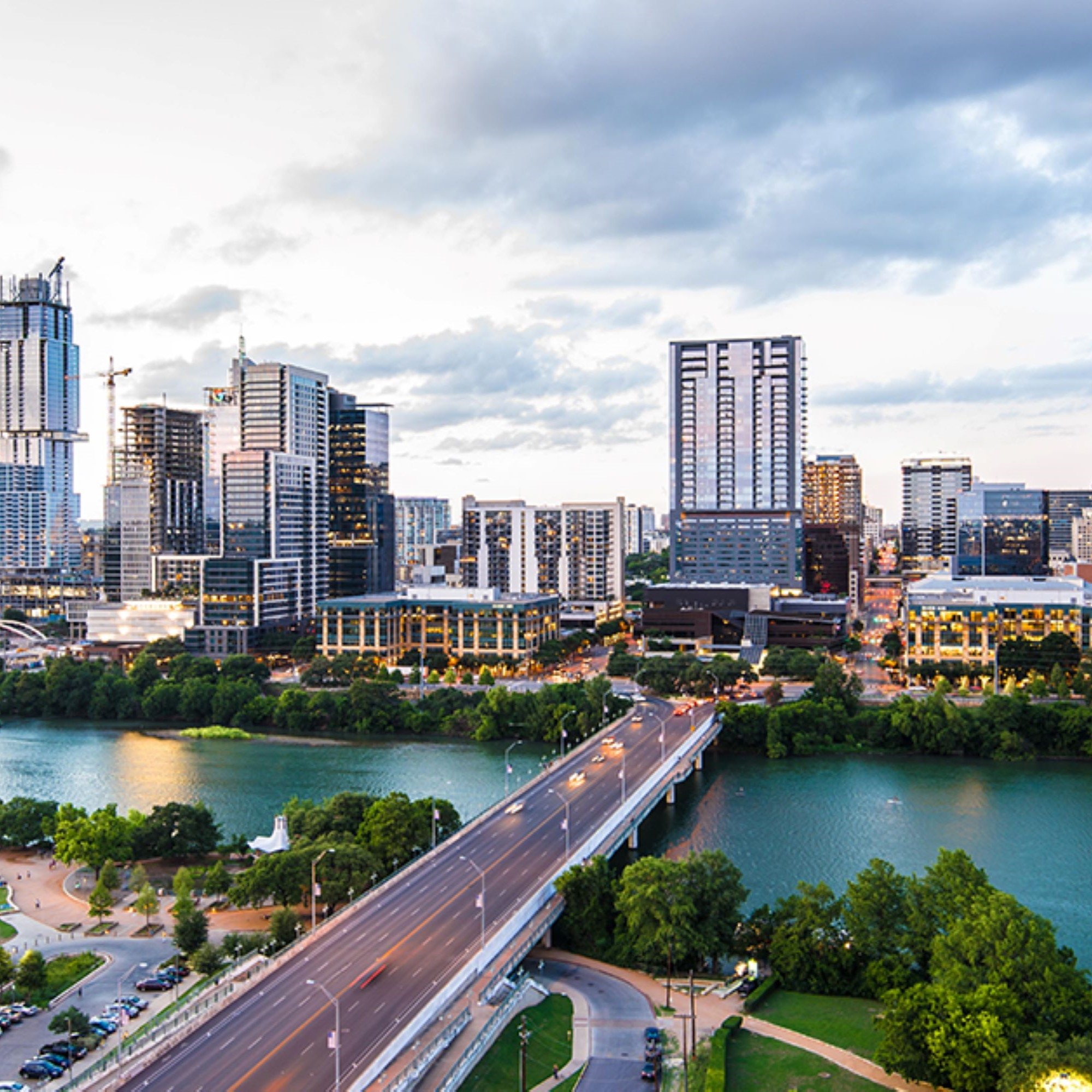 Aerial view of a city skyline with modern high-rise buildings, a bridge crossing a river, and green park areas under a cloudy sky at dusk.