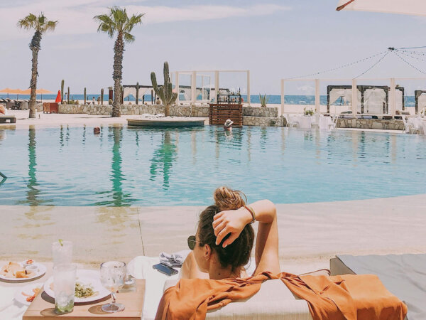 Woman lounging on a poolside chair with a burnt orange cover-up, overlooking an infinity pool with palm trees, a cactus, and the ocean in the background on a sunny day.
