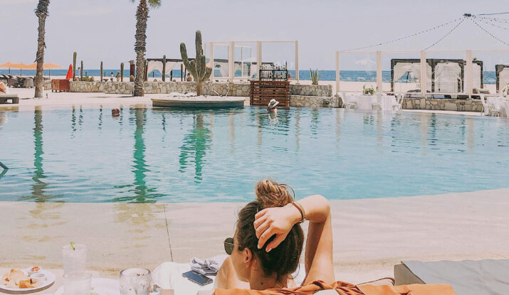 Woman lounging on a poolside chair with a burnt orange cover-up, overlooking an infinity pool with palm trees, a cactus, and the ocean in the background on a sunny day.