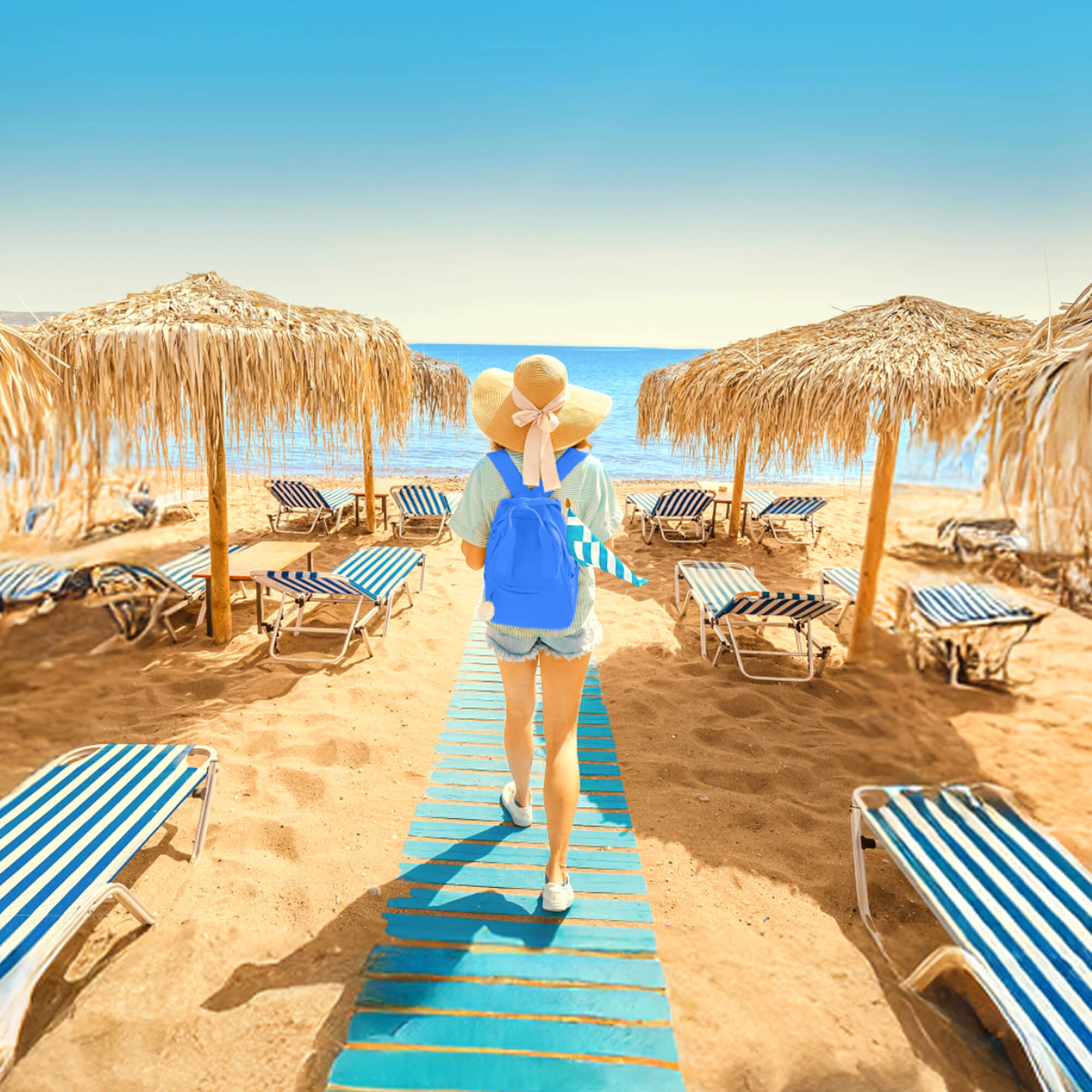 Person wearing a sun hat, light green shirt, denim shorts, and blue backpack walking on a blue wooden pathway through a sandy beach with straw umbrellas and striped lounge chairs, facing the ocean under a clear sky.