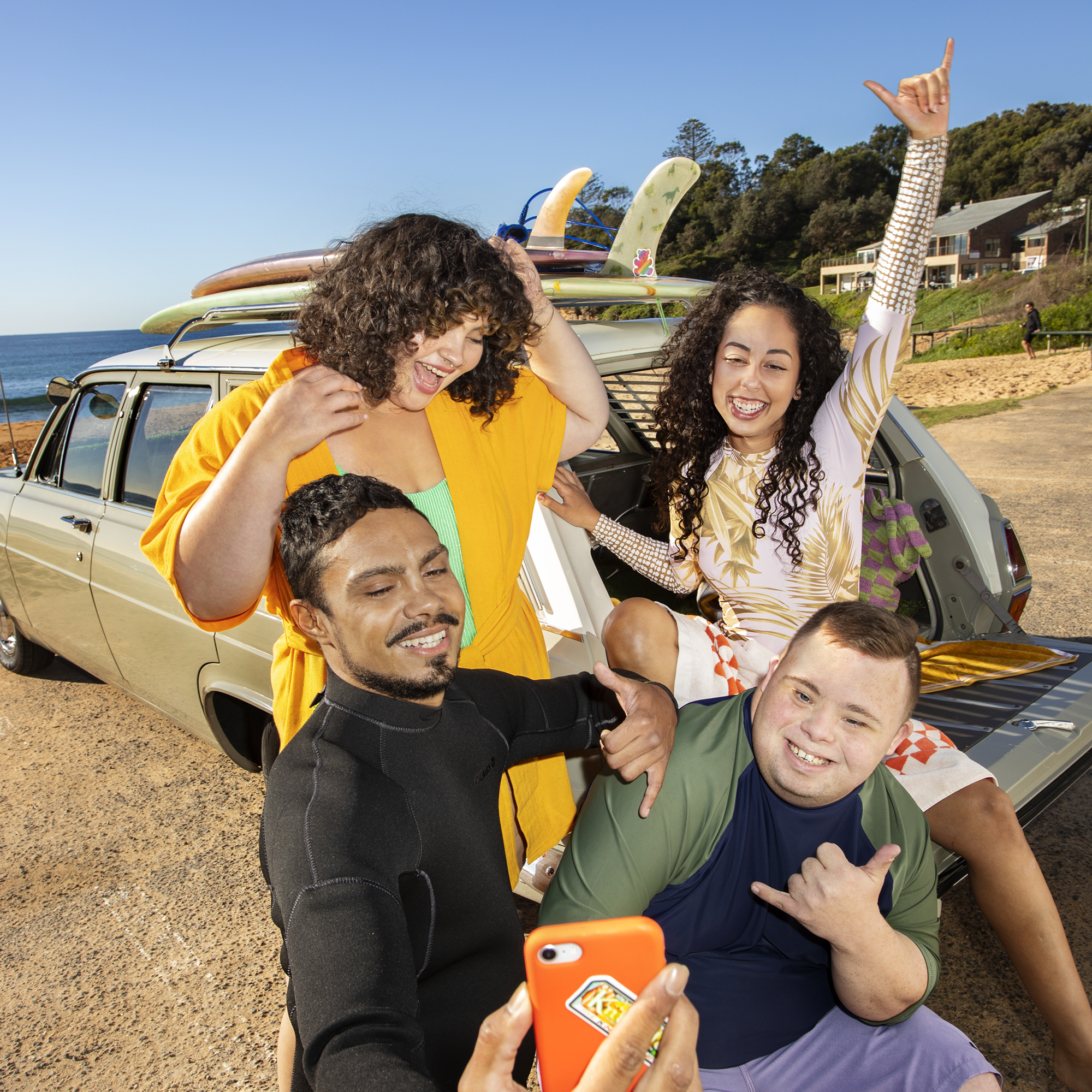 Four friends posing for a selfie near a beach with surfboards on the roof of a vintage car, all smiling and making shaka hand signs, with a clear blue sky and greenery in the background.