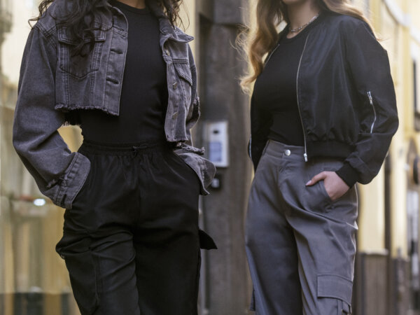 Two women standing confidently on a cobblestone street wearing sunglasses, dark tops, and high-waisted cargo pants; the woman on the left has long curly hair with a cropped denim jacket, and the woman on the right has wavy hair with a black bomber jacket.