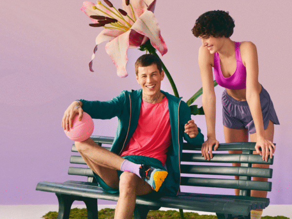 A smiling man in a teal jacket and pink shirt sitting cross-legged on a green bench holding a pink basketball, with a smiling woman in a pink sports bra and purple shorts leaning on the bench beside him, and a large pink flower in the background against a gradient purple and pink backdrop.