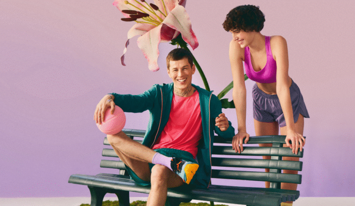 A smiling man in a teal jacket and pink shirt sitting cross-legged on a green bench holding a pink basketball, with a smiling woman in a pink sports bra and purple shorts leaning on the bench beside him, and a large pink flower in the background against a gradient purple and pink backdrop.