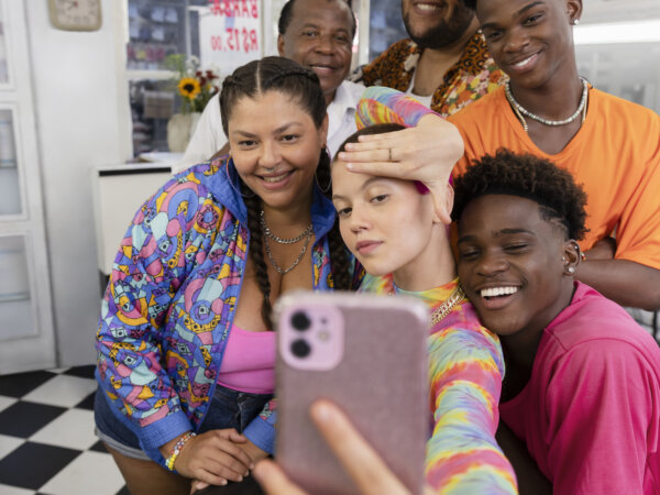 Six diverse friends indoors smiling and posing playfully for a selfie taken by a person sitting in a chair wearing a colorful tie-dye top and checkered shorts, with a checkered black and white floor in the background.