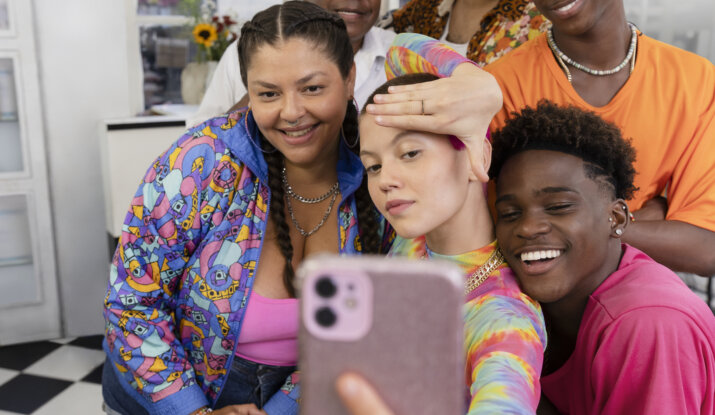 Six diverse friends indoors smiling and posing playfully for a selfie taken by a person sitting in a chair wearing a colorful tie-dye top and checkered shorts, with a checkered black and white floor in the background.