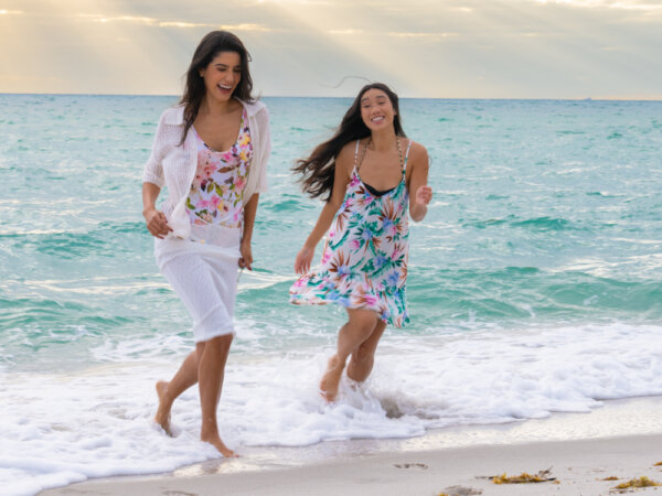 Two women laughing and running barefoot in the shallow ocean waves on a sandy beach with a cloudy sky and sun rays in the background.