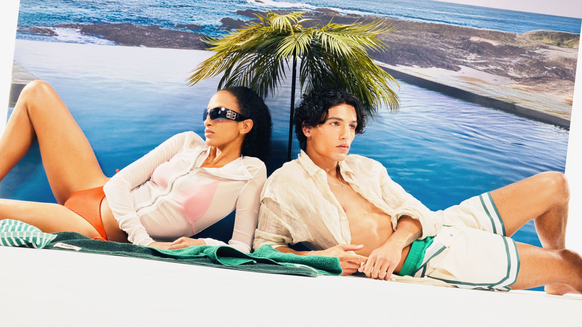A woman in orange bikini bottoms and sunglasses reclining next to a man wearing cream shorts and an open cream shirt, both leaning against a backdrop of a pool and palm tree, lying on green towels.