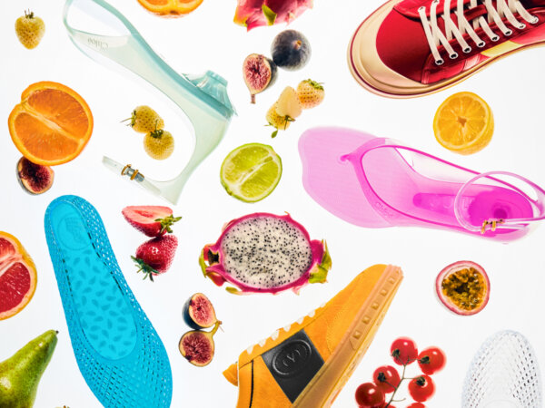 Flat lay of colorful shoes including blue jelly flats, pink jelly sandals, clear jelly heels, and orange sneakers, interspersed with various fresh fruits like dragon fruit, strawberries, figs, oranges, lime, cherry tomatoes, and passion fruit on a white background.