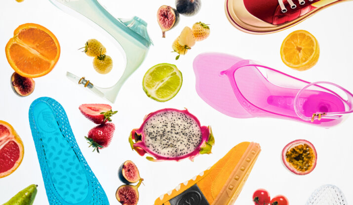 Flat lay of colorful shoes including blue jelly flats, pink jelly sandals, clear jelly heels, and orange sneakers, interspersed with various fresh fruits like dragon fruit, strawberries, figs, oranges, lime, cherry tomatoes, and passion fruit on a white background.