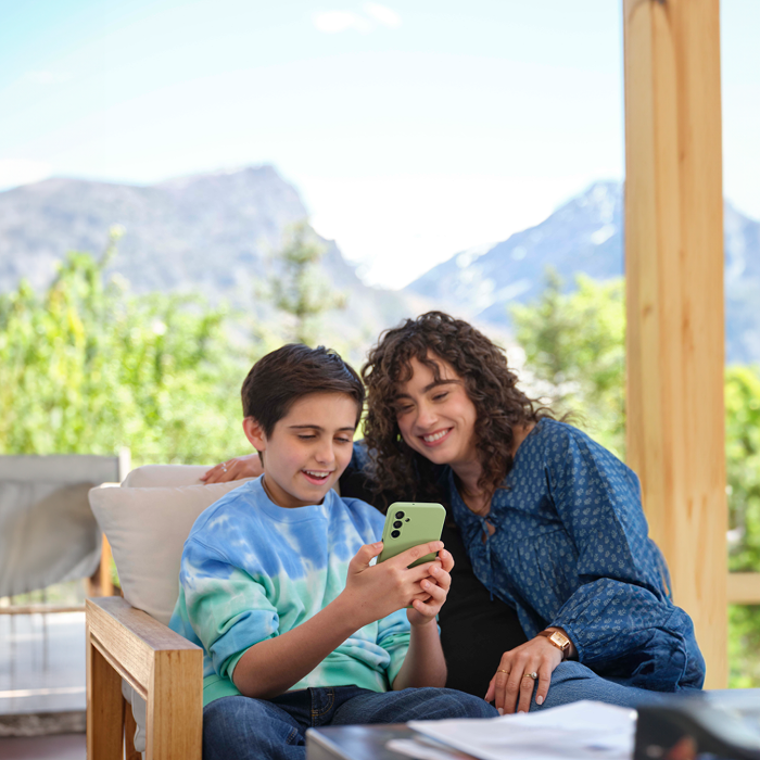 A smiling woman with curly hair and a boy in a blue and green tie-dye sweatshirt sitting on a wooden chair outdoors, looking at a green smartphone together with a blurred mountain and greenery background.