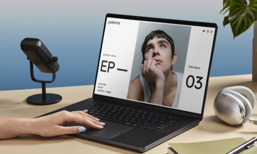 Person with painted nails using a black laptop on a beige desk displaying a podcast webpage featuring a young man resting his face on his hand and looking upward, with a microphone, silver headphones, a green book, and a pen nearby and a green leafy plant in the background.