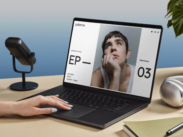 Person with painted nails using a black laptop on a beige desk displaying a podcast webpage featuring a young man resting his face on his hand and looking upward, with a microphone, silver headphones, a green book, and a pen nearby and a green leafy plant in the background.