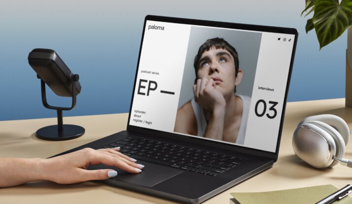 Person with painted nails using a black laptop on a beige desk displaying a podcast webpage featuring a young man resting his face on his hand and looking upward, with a microphone, silver headphones, a green book, and a pen nearby and a green leafy plant in the background.