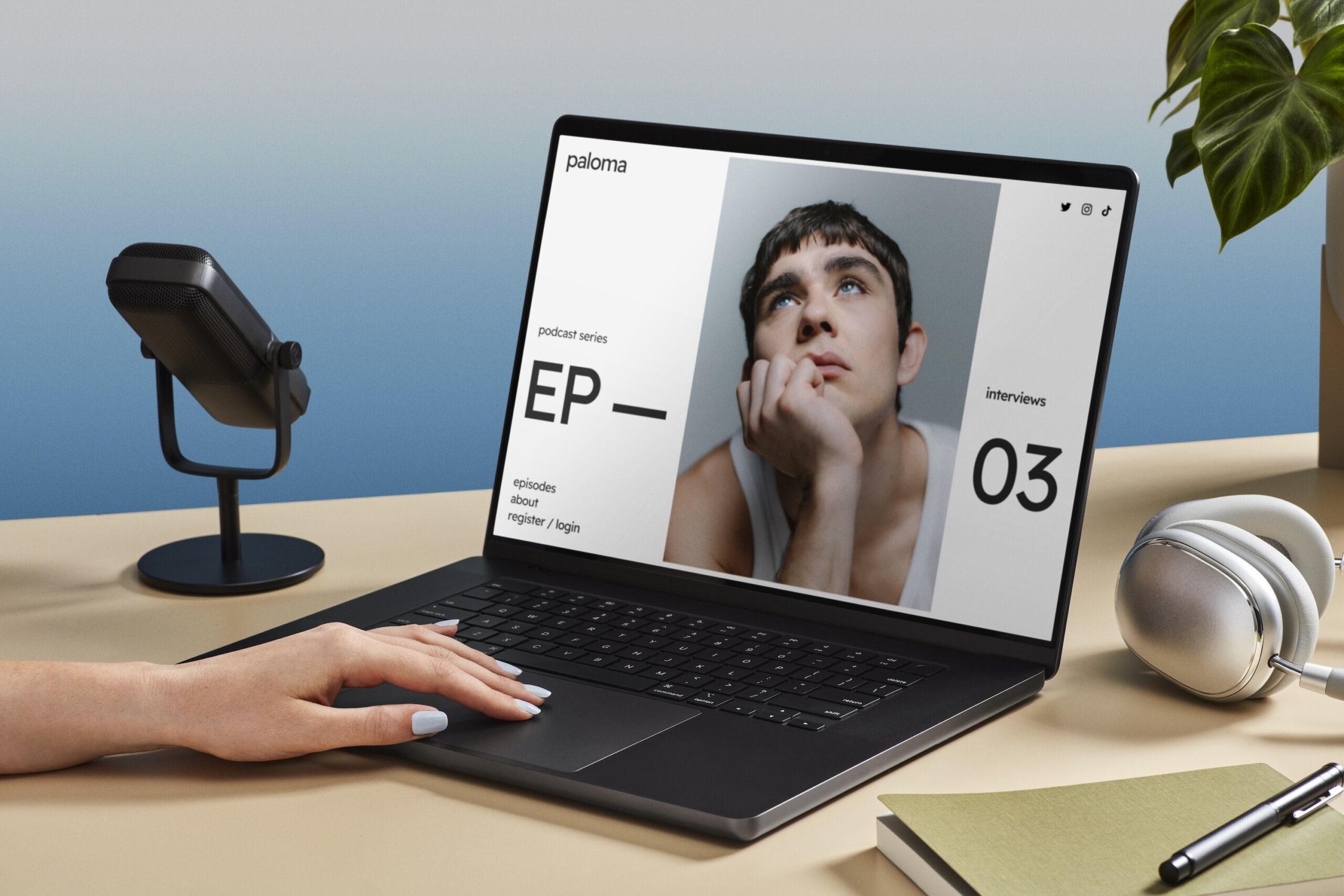 Person with painted nails using a black laptop on a beige desk displaying a podcast webpage featuring a young man resting his face on his hand and looking upward, with a microphone, silver headphones, a green book, and a pen nearby and a green leafy plant in the background.