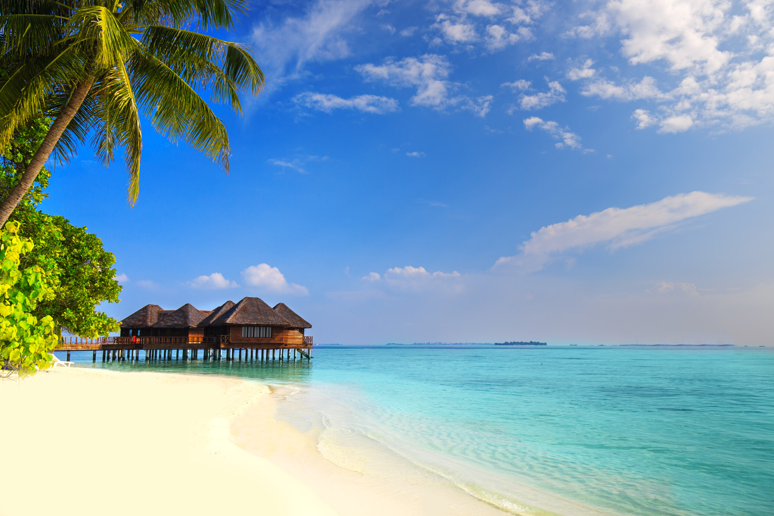 Tropical beach scene with white sand, clear turquoise water, palm trees on the left, and wooden overwater bungalows extending into the sea under a blue sky with scattered clouds.