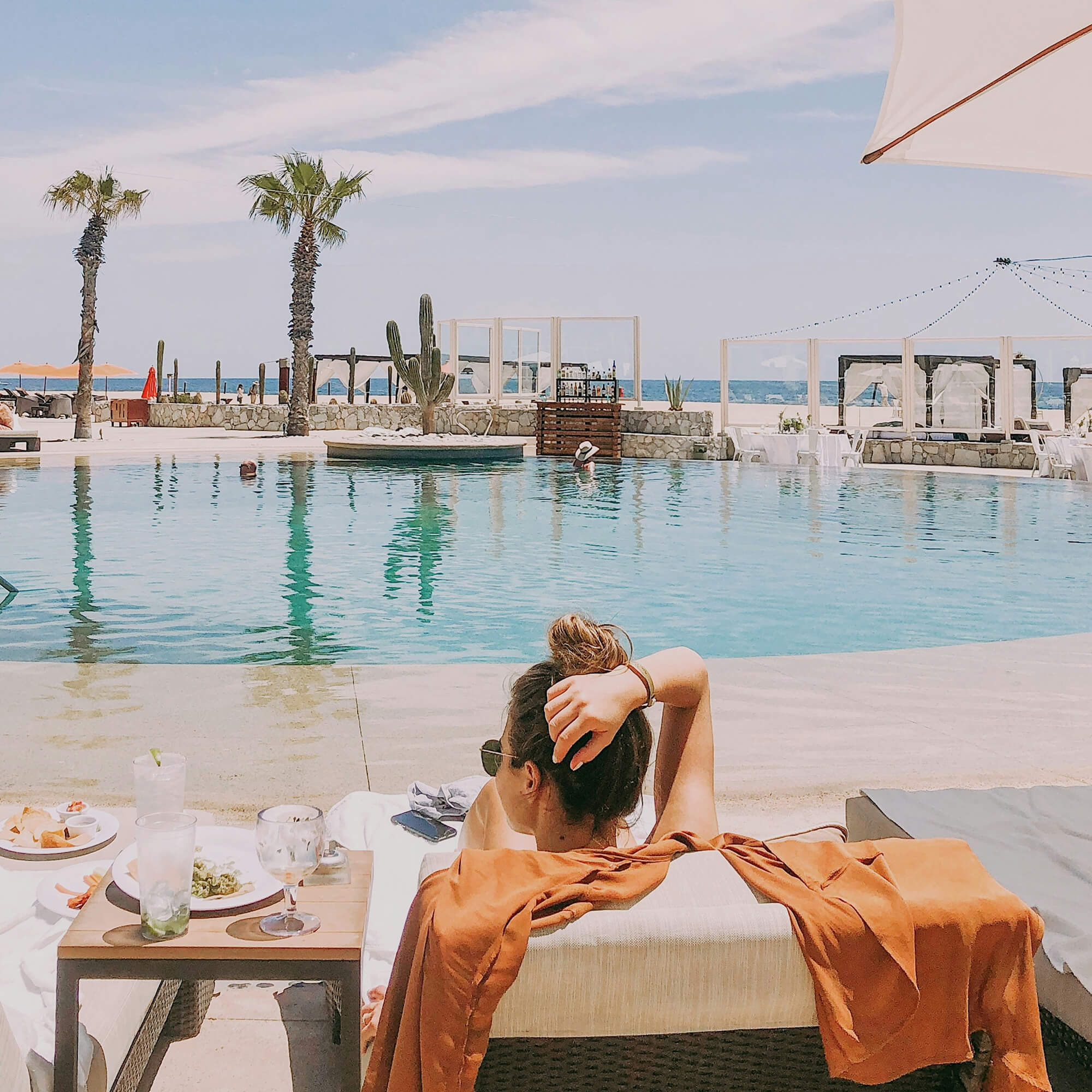 A woman with sunglasses and an orange shirt draped over her lounge chair relaxing by a pool with palm trees, a cactus, and cabanas in the background under a partly cloudy sky.