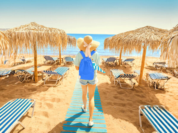 Person wearing a large sun hat and blue backpack walking on a wooden pathway towards the sea on a sandy beach with striped lounge chairs and thatched umbrellas on both sides