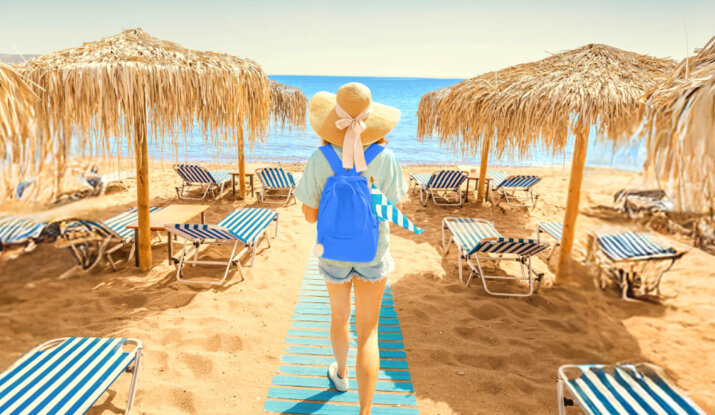 Person wearing a large sun hat and blue backpack walking on a wooden pathway towards the sea on a sandy beach with striped lounge chairs and thatched umbrellas on both sides