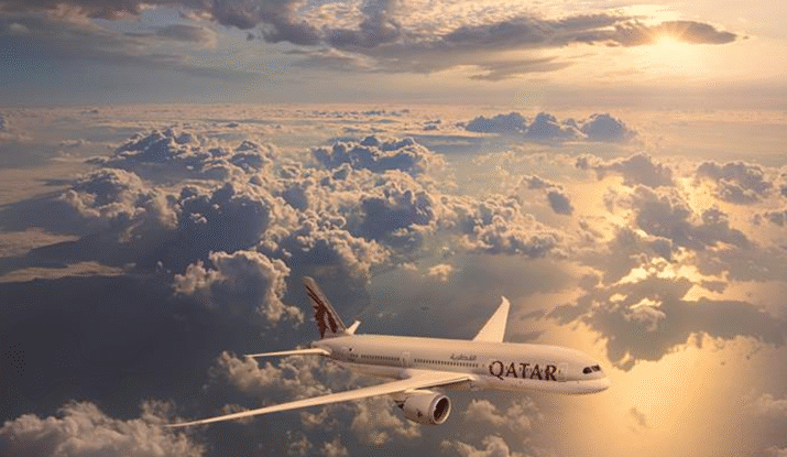 A Qatar Airways plane flying above clouds during a sunset with the sky lit in warm golden tones.