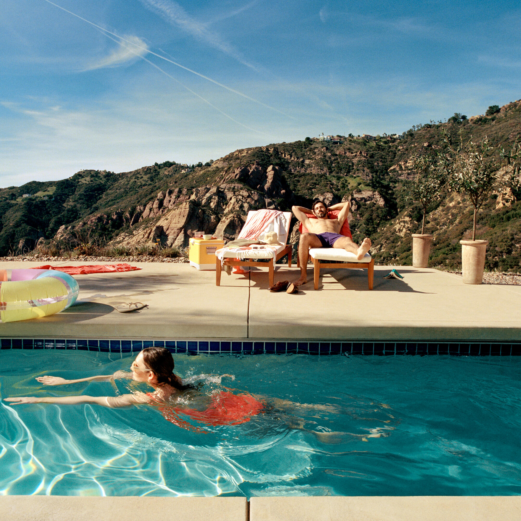 A woman swims in a pool while a man reclines on a lounge chair with hands behind his head in a sunny outdoor setting with mountains in the background and two potted plants nearby.